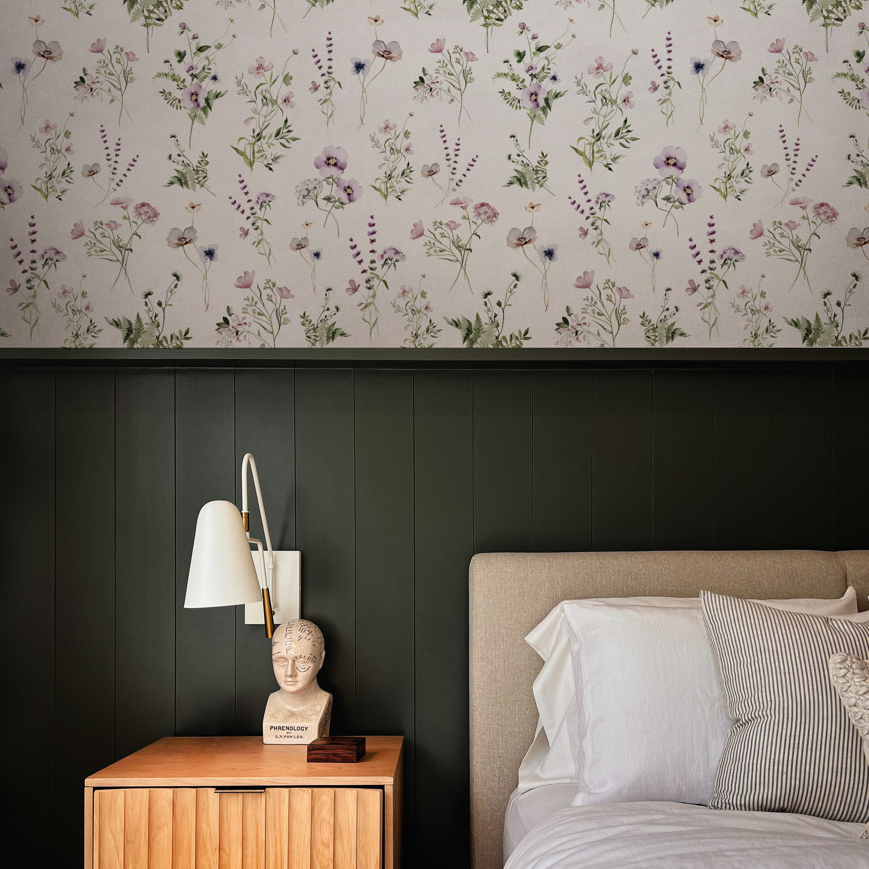 A section of a bedroom showing the 'Midsummer Watercolour Bouquet Wallpaper - Raspberry' with delicate watercolor wildflowers in shades of purple and green, above a dark green wainscoting. Beside the bed, a modern lamp illuminates a wooden nightstand featuring a phrenology head decor piece, contributing to the room's chic and eclectic style