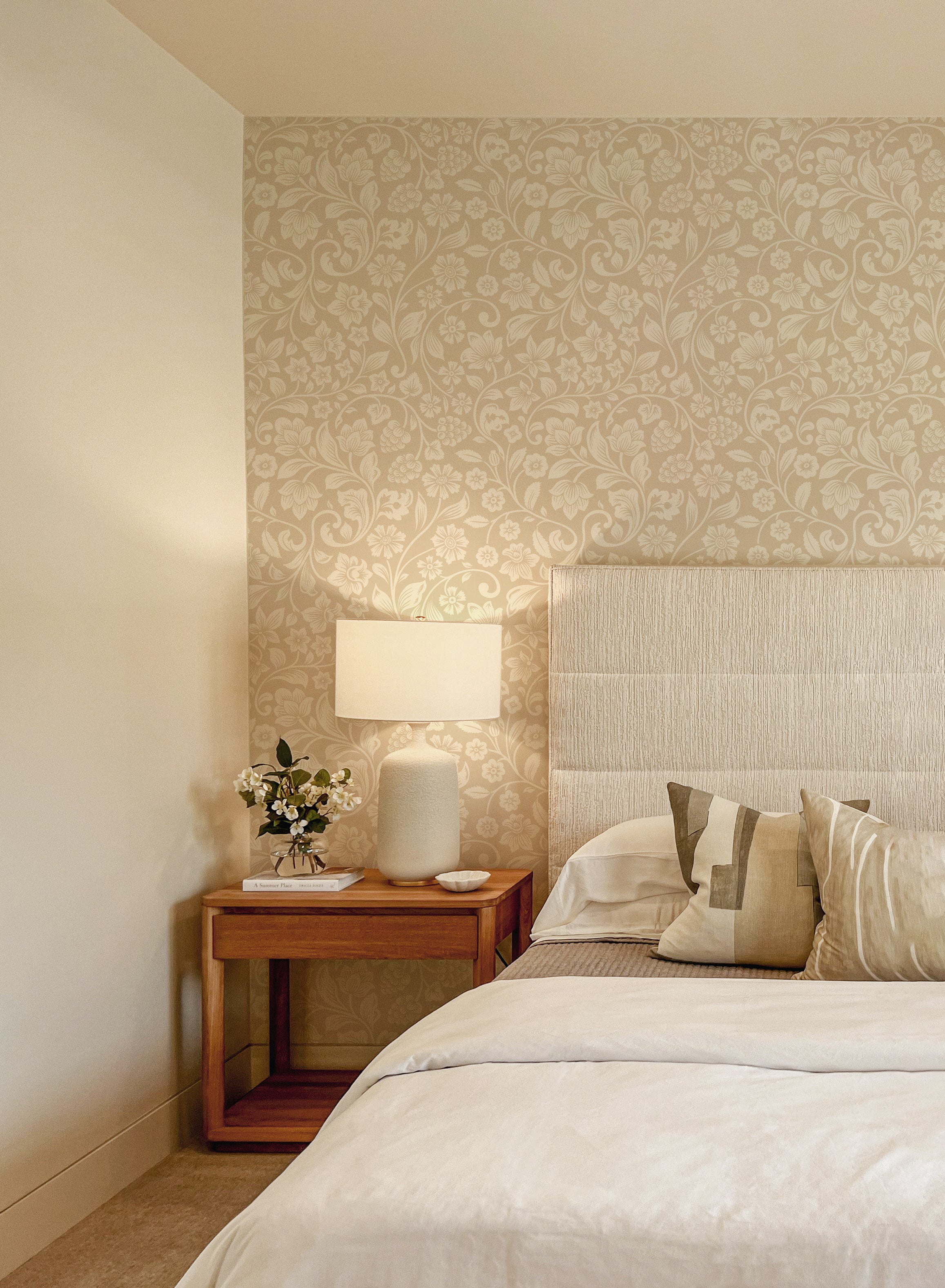 A bedroom wall covered in 'Vintage Floral Wallpaper' creates a warm and inviting ambiance, complementing the neutral bedding and wooden bedside table, with a white ceramic lamp adding to the room's serene decor.