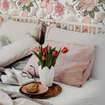 A cozy bedroom setting with the Floral Wallpaper - Berry Pink, featuring large berry-pink roses and various pink-toned flowers creating a vibrant and romantic botanical scene behind a bed adorned with soft linens and a vase of red tulips.