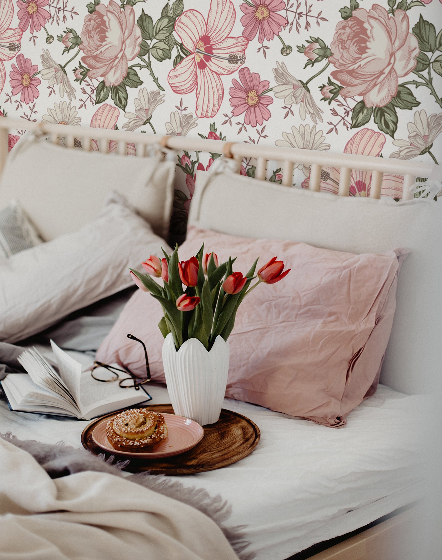 A cozy bedroom setting with the Floral Wallpaper - Berry Pink, featuring large berry-pink roses and various pink-toned flowers creating a vibrant and romantic botanical scene behind a bed adorned with soft linens and a vase of red tulips.