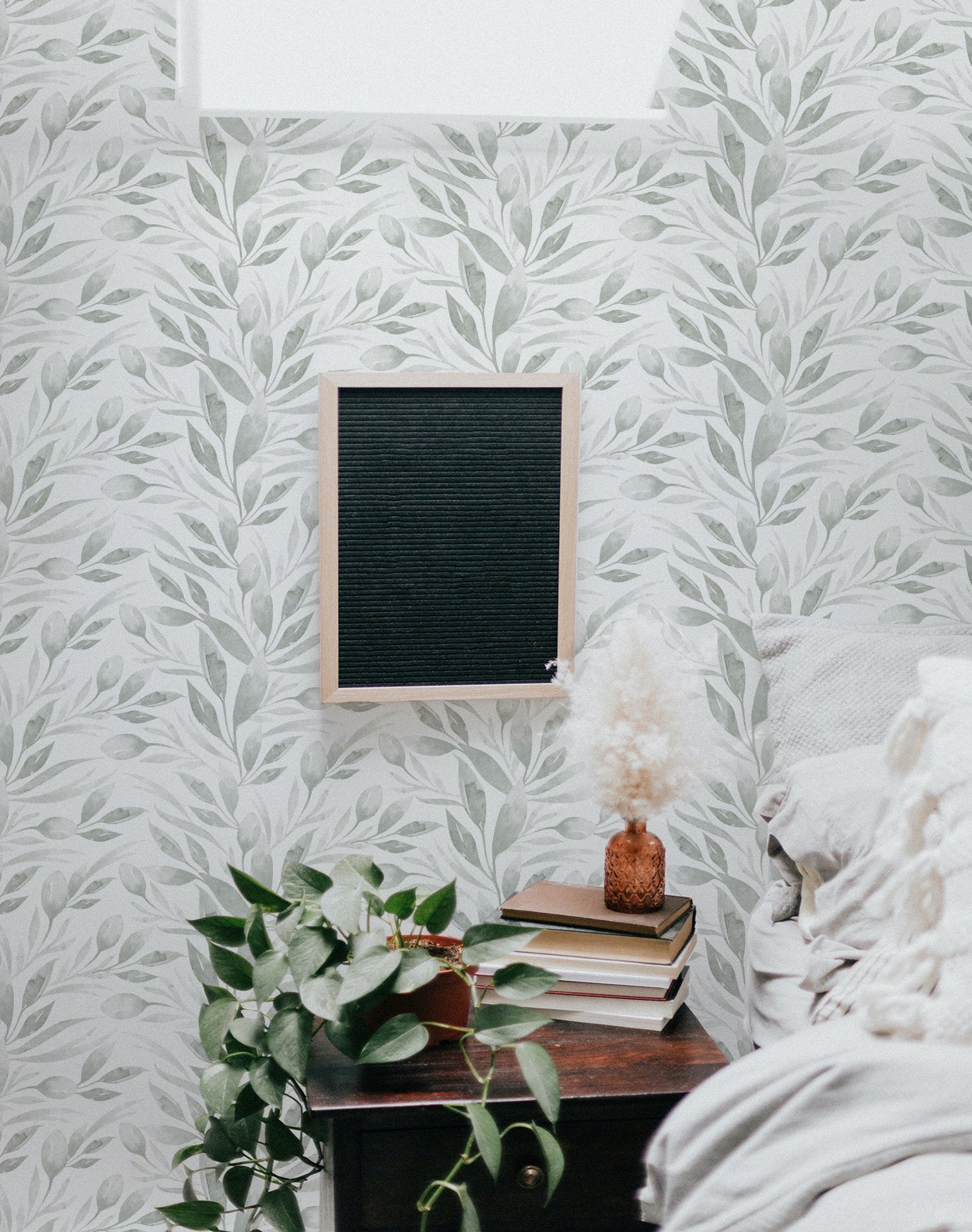 A cozy corner of a room featuring Watercolour Spring Leaf Wallpaper, which displays soft gray and green leaf patterns on a light background, paired with indoor plants and vintage furniture for a serene ambiance