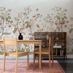 displays a home office area enhanced by the same Vintage Floral Mural, exuding a tranquil and inspirational workspace. The mural's detailed flowers and fauna span across the wall, infusing the room with elegance and a touch of nature. An oak wood desk and chair sit before the wall, accompanied by a bookshelf filled with assorted decorative objects and books, complemented by soft, pink-toned textiles that echo the colors in the wallpaper.