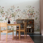 displays a home office area enhanced by the same Vintage Floral Mural, exuding a tranquil and inspirational workspace. The mural's detailed flowers and fauna span across the wall, infusing the room with elegance and a touch of nature. An oak wood desk and chair sit before the wall, accompanied by a bookshelf filled with assorted decorative objects and books, complemented by soft, pink-toned textiles that echo the colors in the wallpaper.
