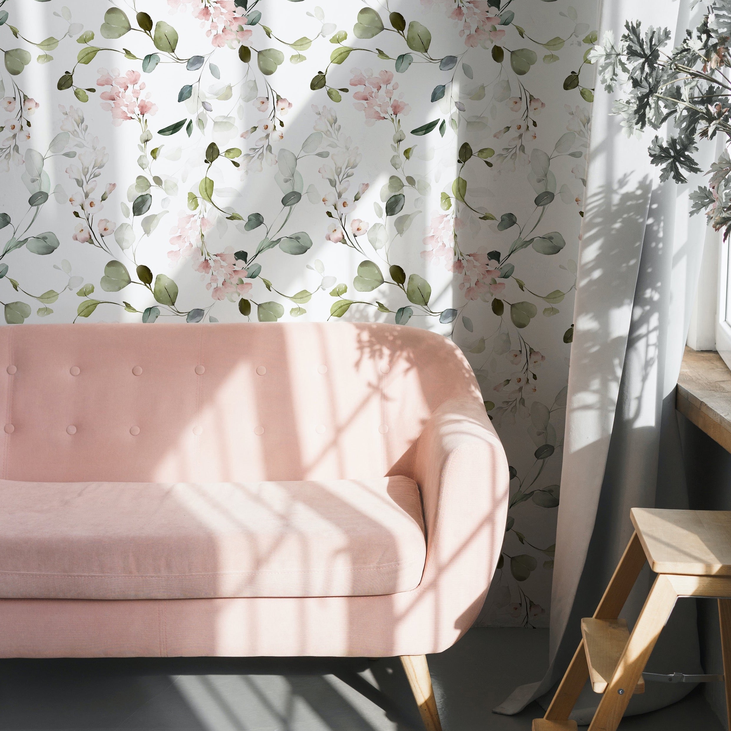 A chic living space illuminated by natural light, highlighting the Pink Floral & Herbs Wallpaper, adorned with blush pink florals and greenery. The wall art complements a plush, pastel pink tufted sofa and a minimalist wooden stool, evoking a fresh, botanical ambiance.