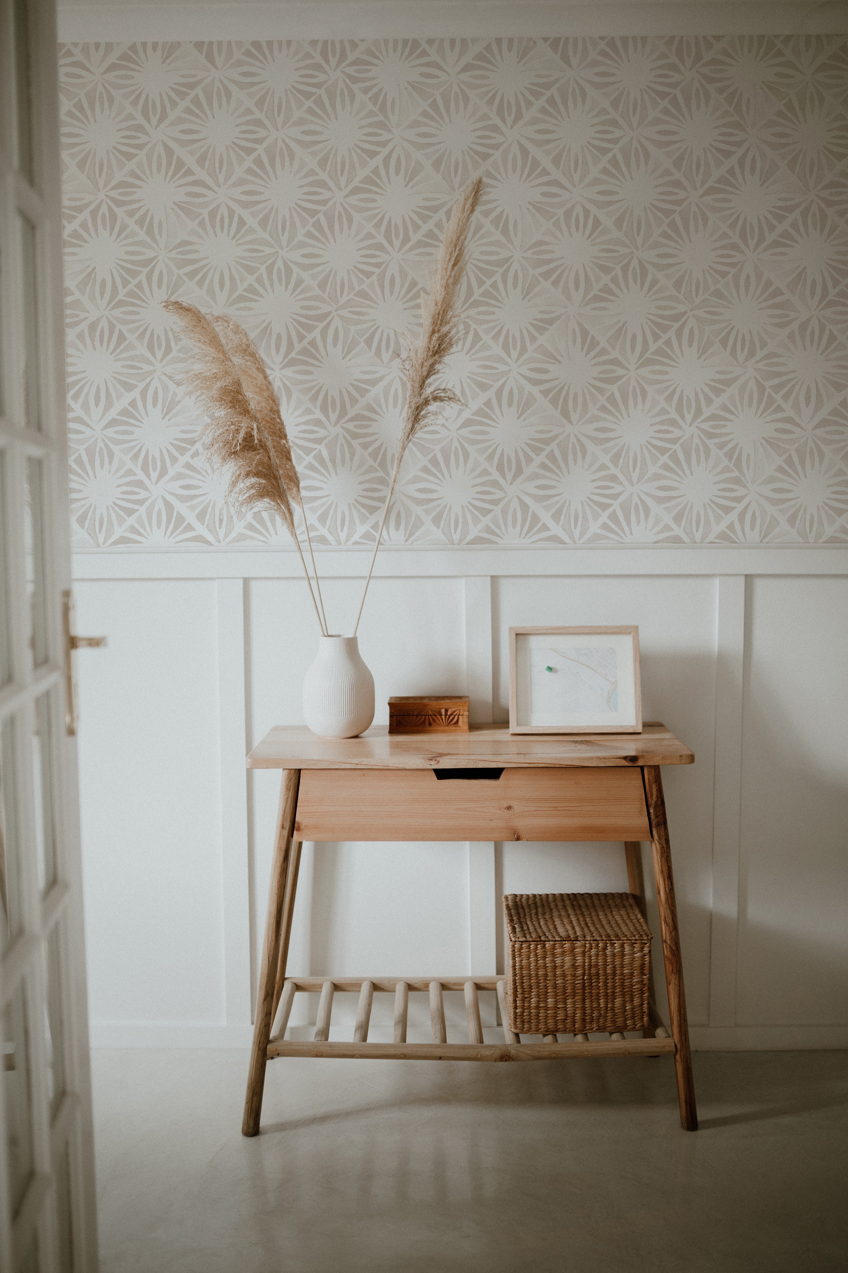  serene workspace with Moroccan Tile Wallpaper II - Linen, showcasing a soft beige geometric pattern, complemented by a natural wooden desk, a textured vase with pampas grass, and simplistic framed artwork, creating a warm and inviting atmosphere.