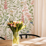 A well-lit dining area with a warm wooden table casting a long shadow over the floor, set against a wall covered with the Watercolour Floral and Leaf Wallpaper. A clear vase with fresh tulips sits on the table, complementing the wallpaper's floral design and creating a harmonious blend of real and illustrated nature