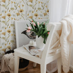 A cozy corner featuring the Watercolour Sunflower Wallpaper adorned with charming sunflowers and delicate flora in watercolor tones of yellow and green. A white chair with a vase of vibrant tulips adds a fresh touch to the inviting scene.