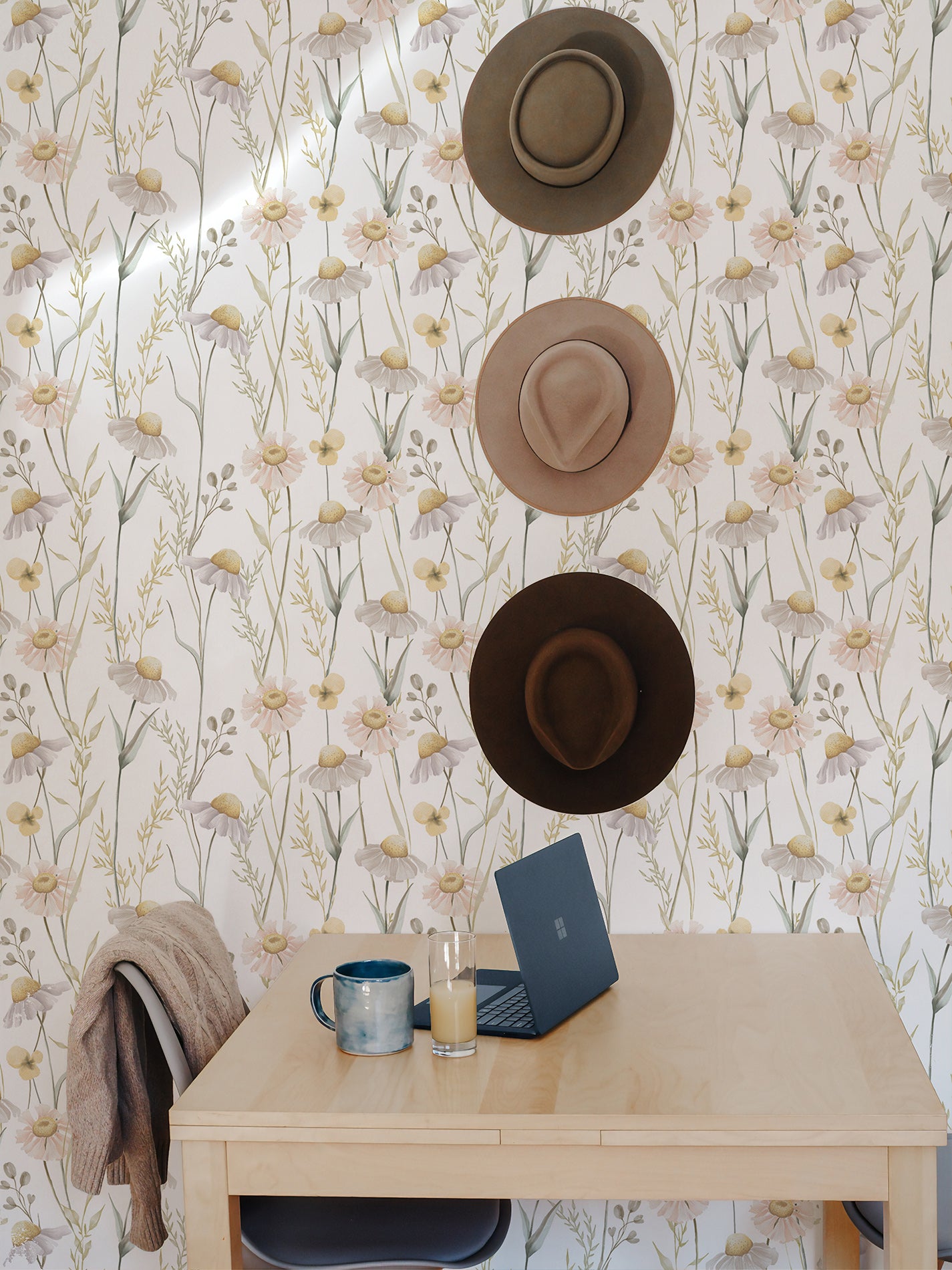 A creative workspace with Watercolour Daisy Wallpaper providing a refreshing and artistic backdrop. The wall is adorned with various hats, suggesting a personal touch, while a simple desk with a laptop, a mug, and a draped sweater add to the lived-in warmth of the space.
