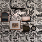 A close-up view of a wall decorated with the "Neutral Geometric Wallpaper I," displaying its intricate and dynamic black geometric designs against a crisp white backdrop. The wallpaper serves as a bold backdrop for various art pieces and sculptures, enhancing the artistic vibe of the space.