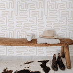A rustic yet chic setting featuring a sturdy wooden bench against a playful, ecru geometric wallpaper background. On the bench, a neat stack consisting of a cozy gray sweater, a beige felt hat, and a speckled mug suggests a moment of leisure and style.