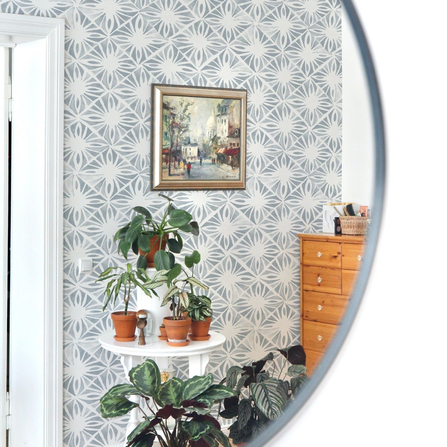 A room corner with a white side table hosting potted plants is reflected in a circular mirror, set against the Moroccan Tile II Wallpaper, which showcases an intricate white and pale blue geometric pattern for an elegant and airy feel.