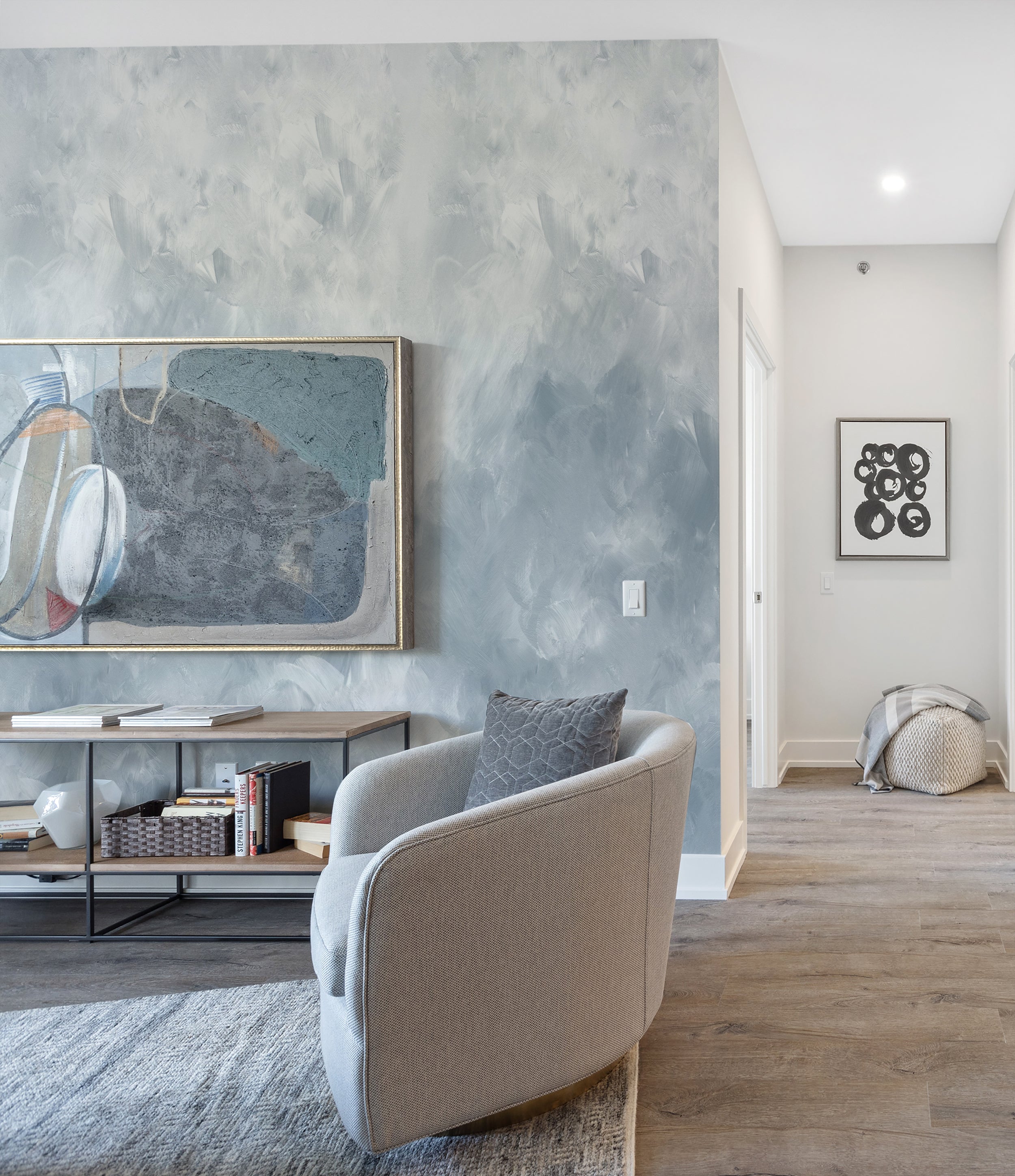 A contemporary living room corner with a curvy light grey armchair and a textured pale blue brush stroke wallpaper creating an abstract backdrop. The room is styled with a modern artwork and a sleek black console table, embodying a chic and artistic ambiance.
