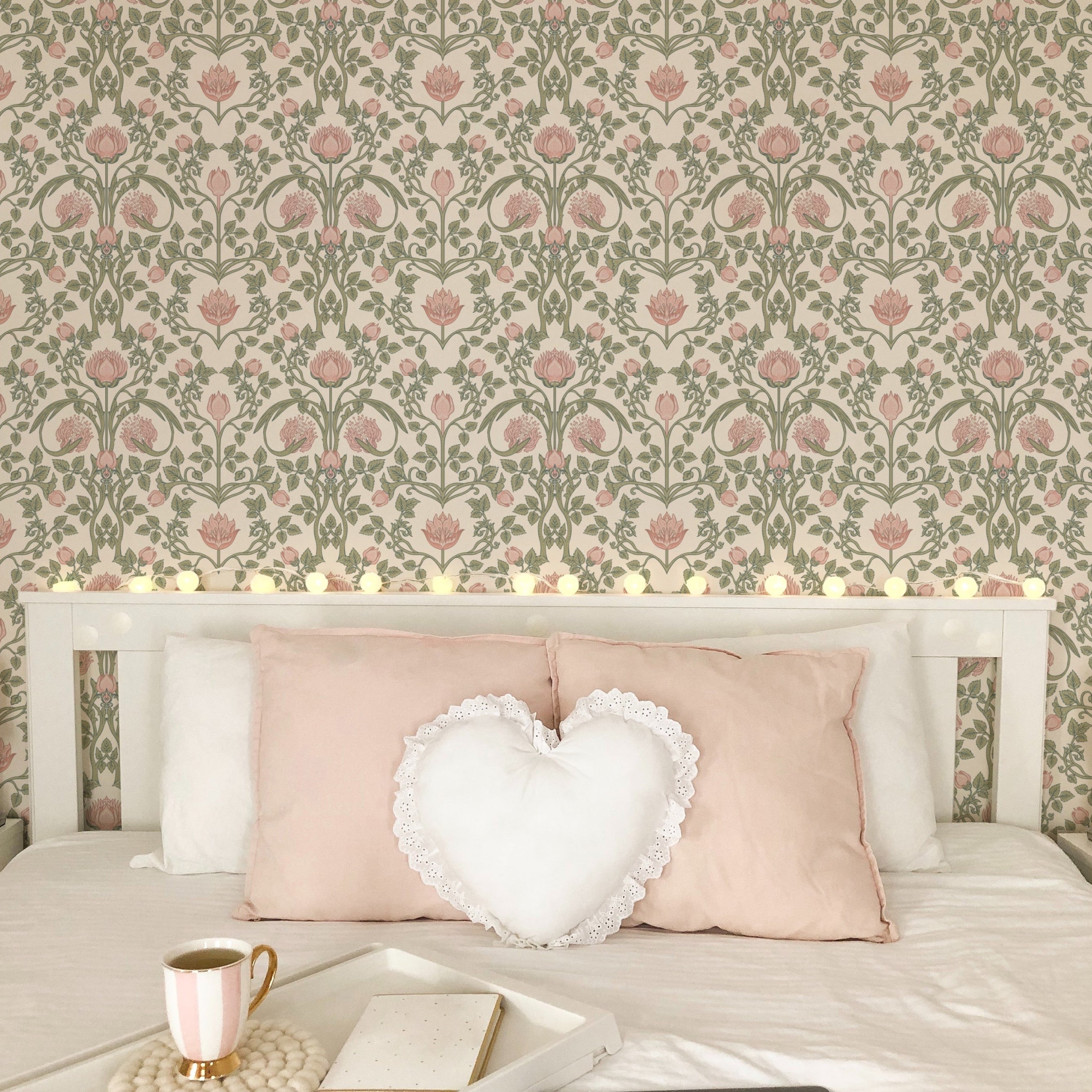A serene bedroom setting with a white bed adorned with soft pink pillows, a heart-shaped cushion, and a serving tray, complemented by the pastel floral damask wallpaper in the background