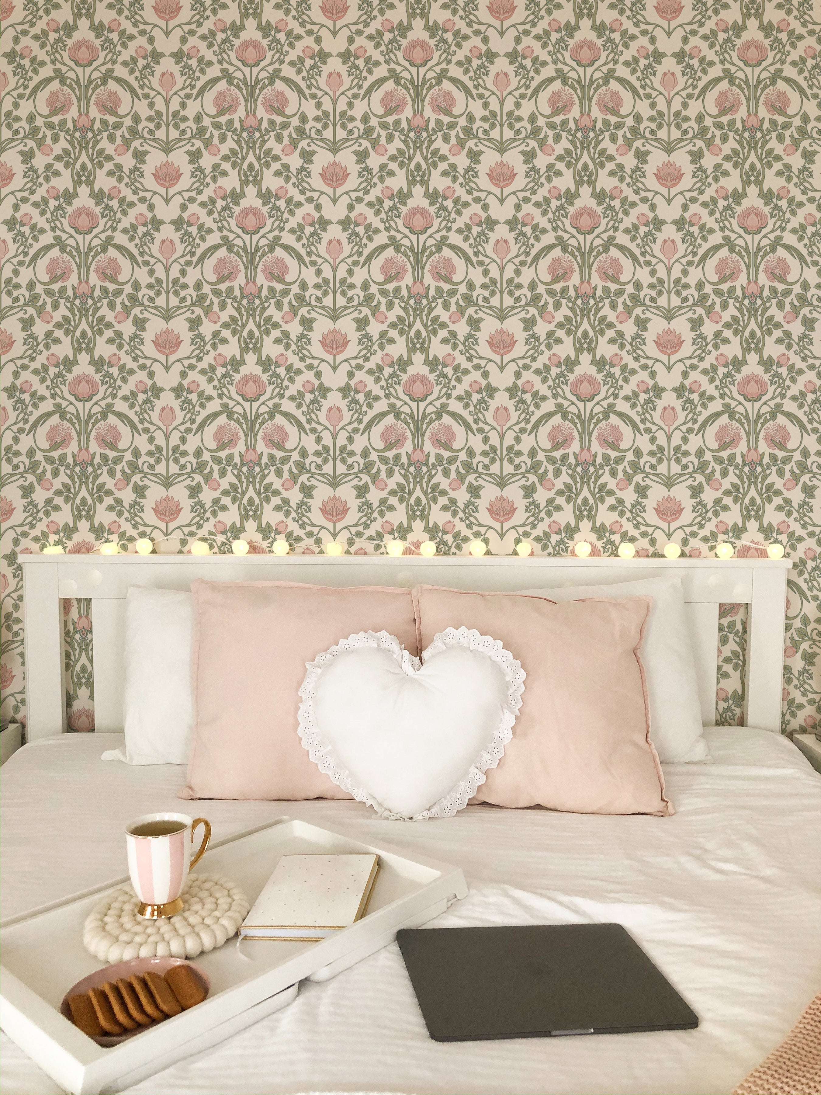 A romantic bedroom ambiance with glowing string lights over a bed, plush pink pillows, a heart-shaped cushion, and the pastel floral damask wallpaper creating a dreamy space