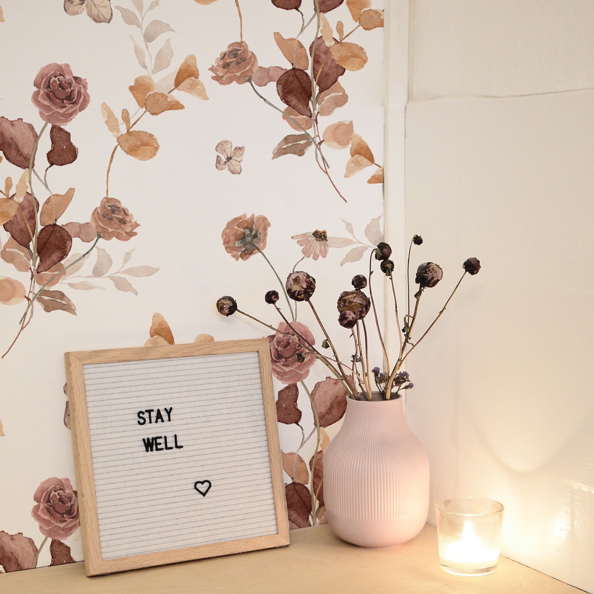 A cozy home setup featuring Autumn Floral Wallpaper with a small wooden sign that reads 'STAY WELL', complemented by a vase of dried flowers and a softly lit candle, emphasizing the wallpaper's soothing floral aesthetic.