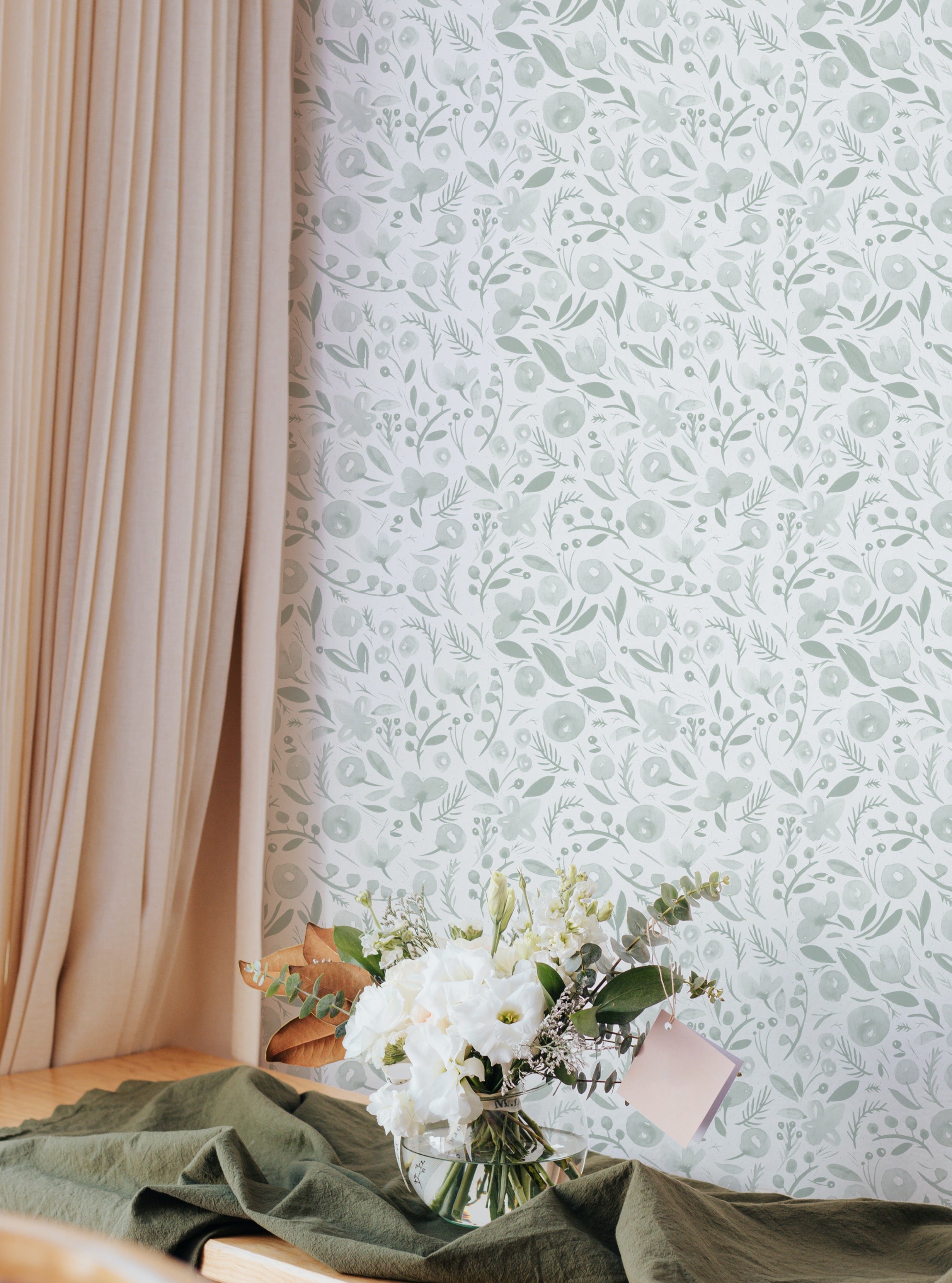 A serene corner setup featuring the Botanica Wallpaper, which displays a delicate botanical pattern of light green foliage and small blooms on a soft white background. This wallpaper adds a fresh, airy feel to the space, complemented by a light floral arrangement and pastel-colored drapery.