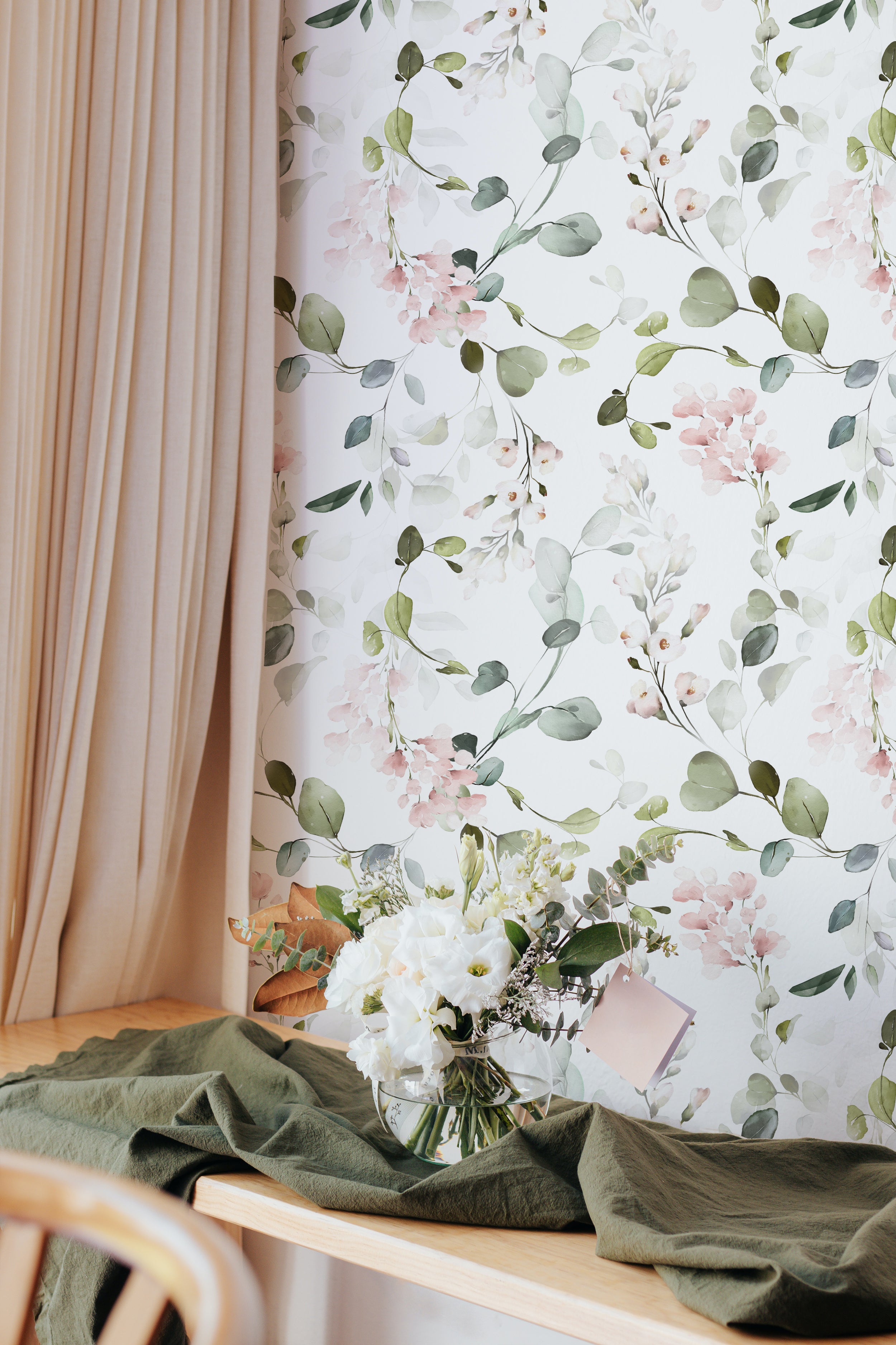A corner of a bright room showcasing the Pink Floral & Herbs Wallpaper, paired with a wooden table dressed with a sage green throw and a bouquet of white flowers. The graceful floral pattern exudes a calm and sophisticated atmosphere.
