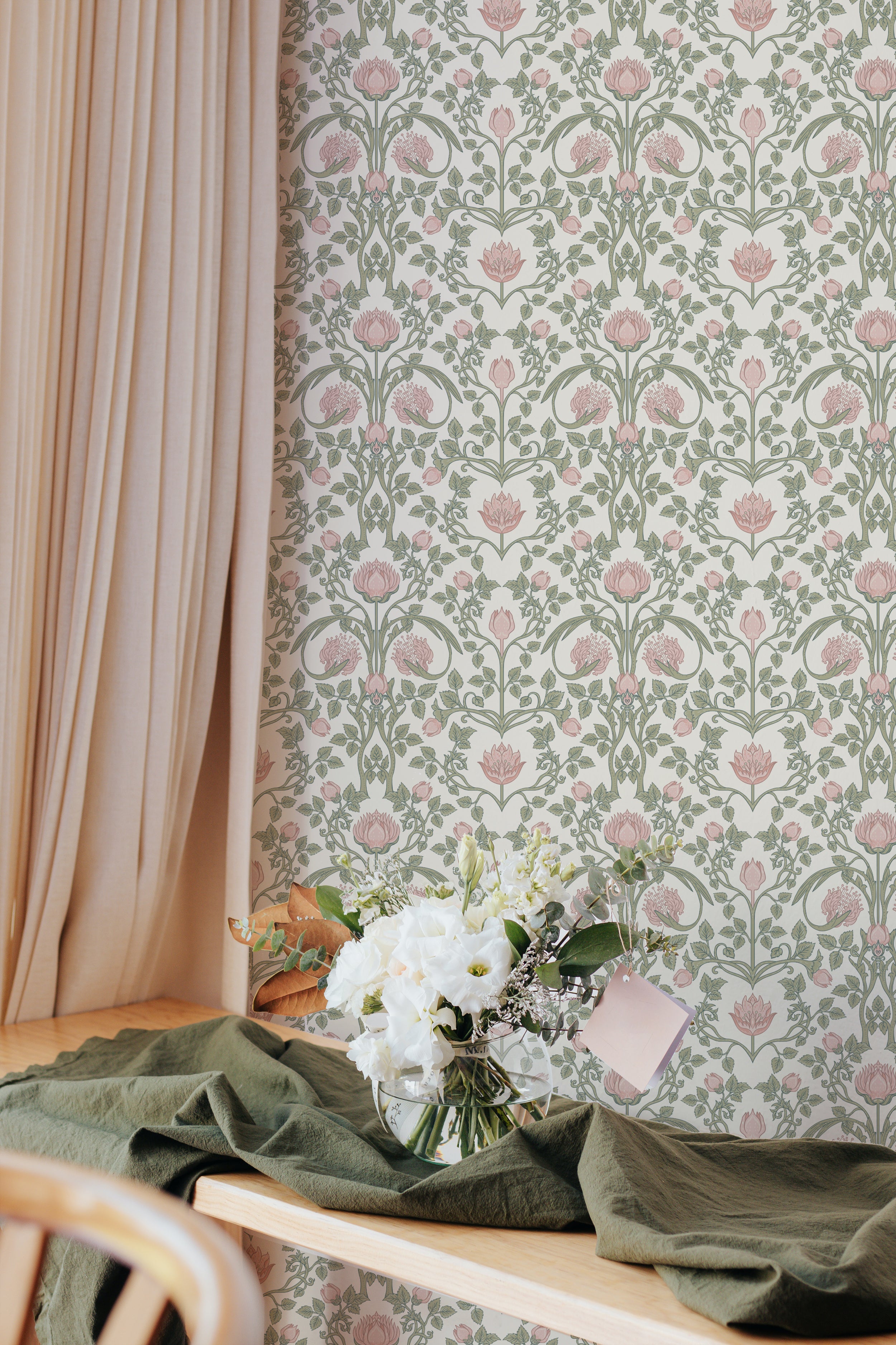 A charming dining area with a wooden table draped with a green cloth, a bouquet of white flowers, and soft drapery, against a wall with pastel floral damask wallpaper.