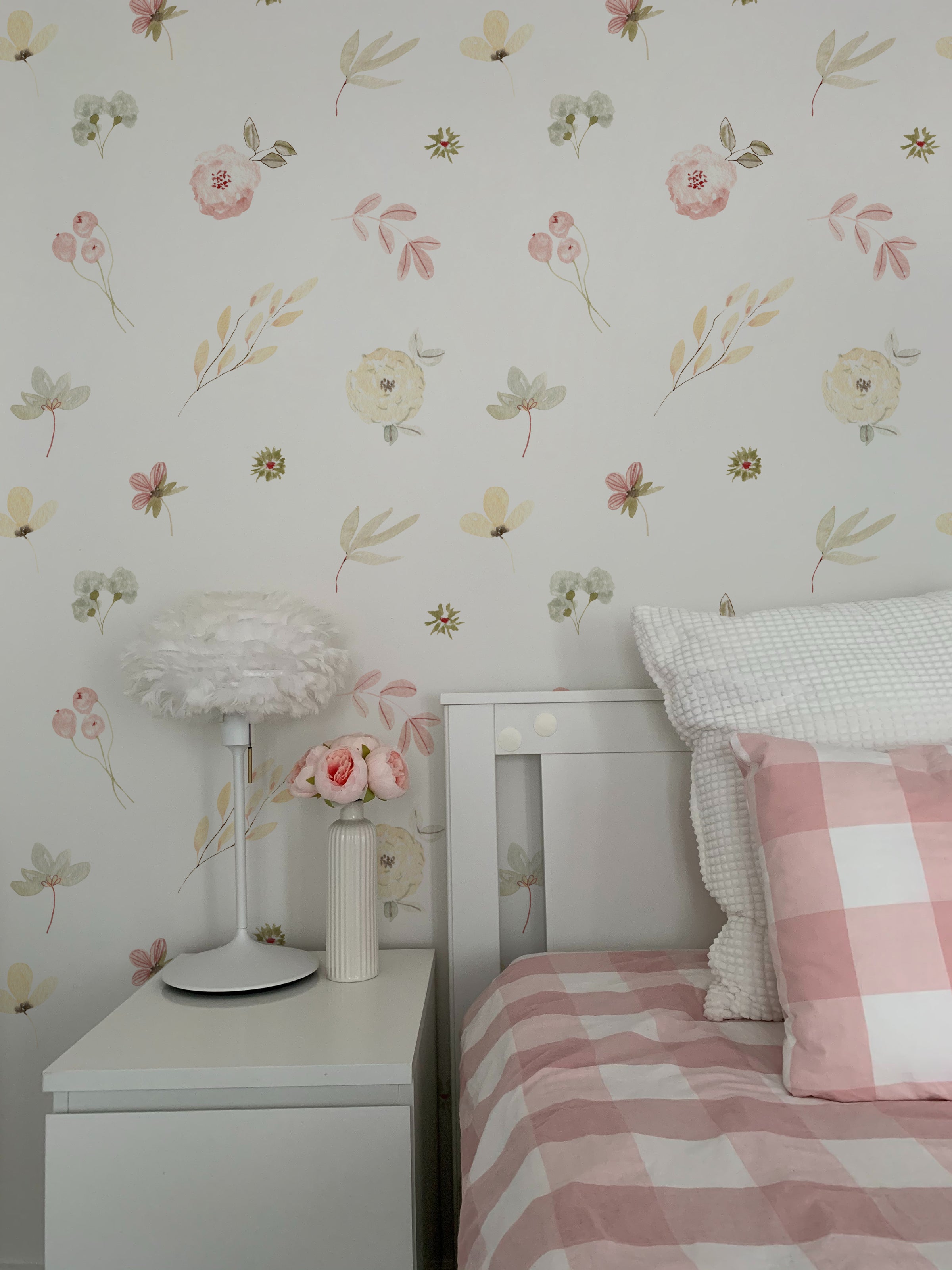 A charming bedroom setup highlights the Pink Watercolour Floral Wallpaper as a beautiful accent. The floral design creates a soothing atmosphere, paired with a cozy bed with pink and white bedding, a feathered lampshade, and a simple white nightstand adorned with fresh flowers for a touch of elegance.