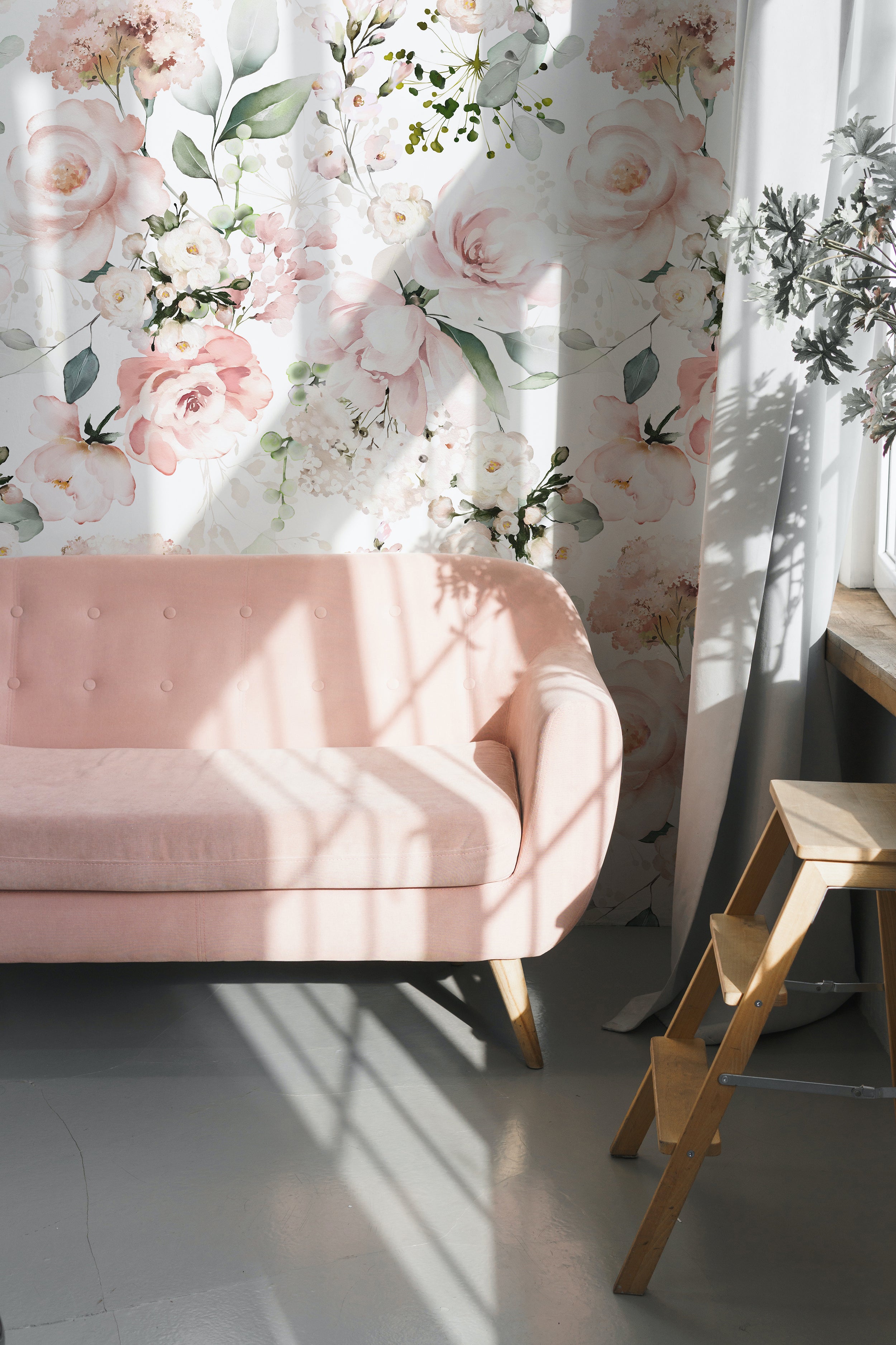 A chic living space accented with the Pink Floral and Herbs Wallpaper - 50", which adorns the wall with its lush, watercolor-style blooms and foliage. A plush pink sofa is positioned against the wallpaper, basking in the natural light that filters through a window, highlighting the soft, pastel colors of the floral pattern.