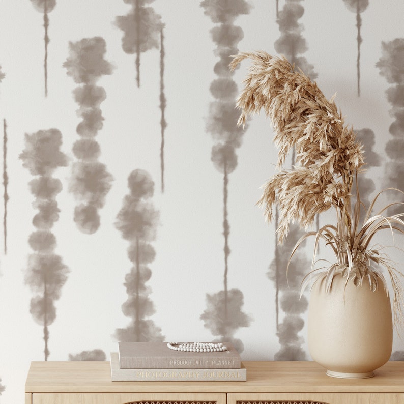 A chic corner of a room showcases the 'Shibori Watercolour Wallpaper', with a unique watercolor tie-dye pattern in neutral tones creating vertical stripes, beside a natural wood sideboard and a tall vase with pampas grass, evoking a tranquil, contemporary vibe.