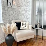 A comfortable sitting area featuring a beige armchair, a small round black side table, and a vase with pampas grass. The white wallpaper with black floral design enhances the room's minimalist aesthetic.