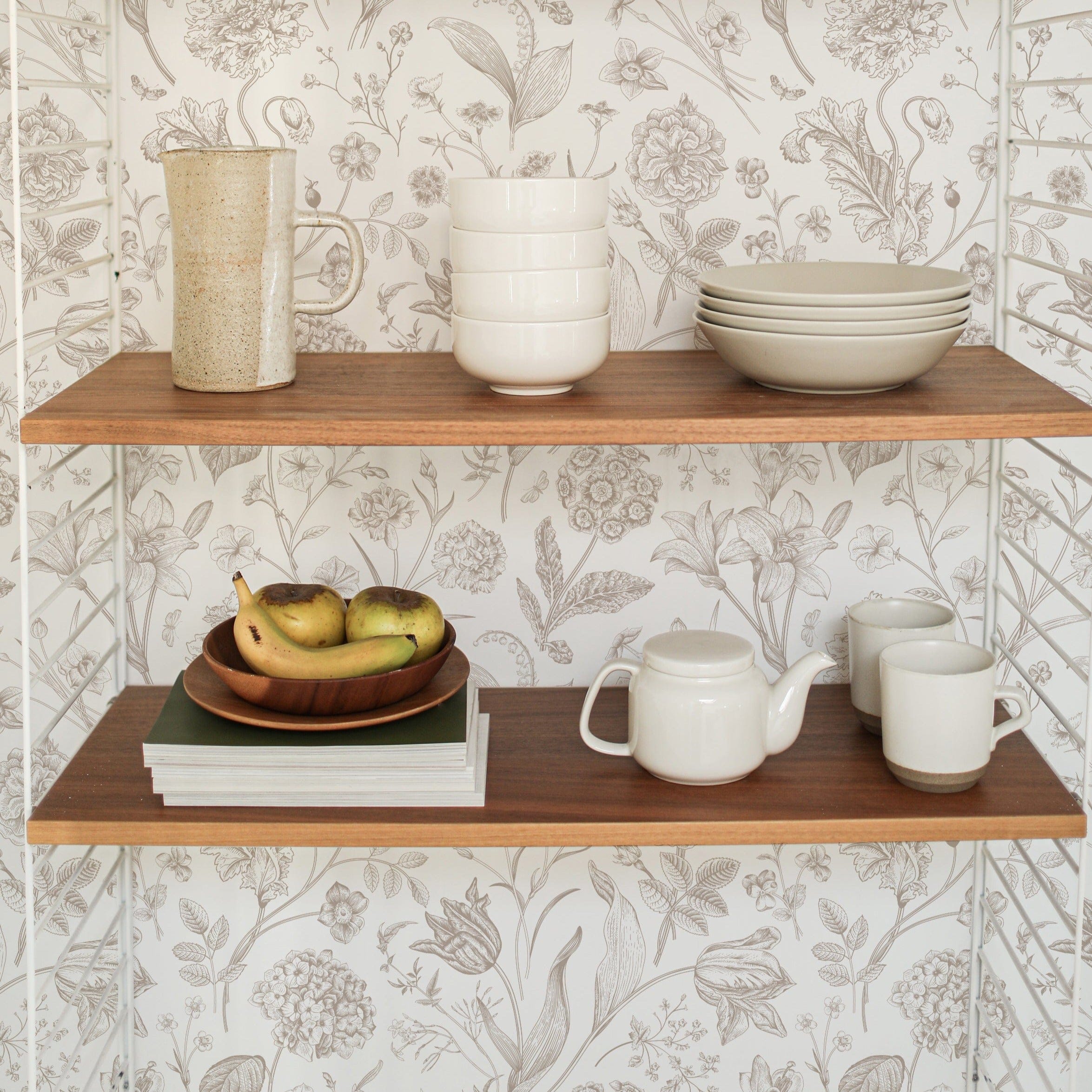 Open wooden shelves against a wall covered with Toile De Jouy Floral Wallpaper, which is adorned with delicate floral drawings. The shelves hold an array of kitchenware, adding a functional touch to the decorative space