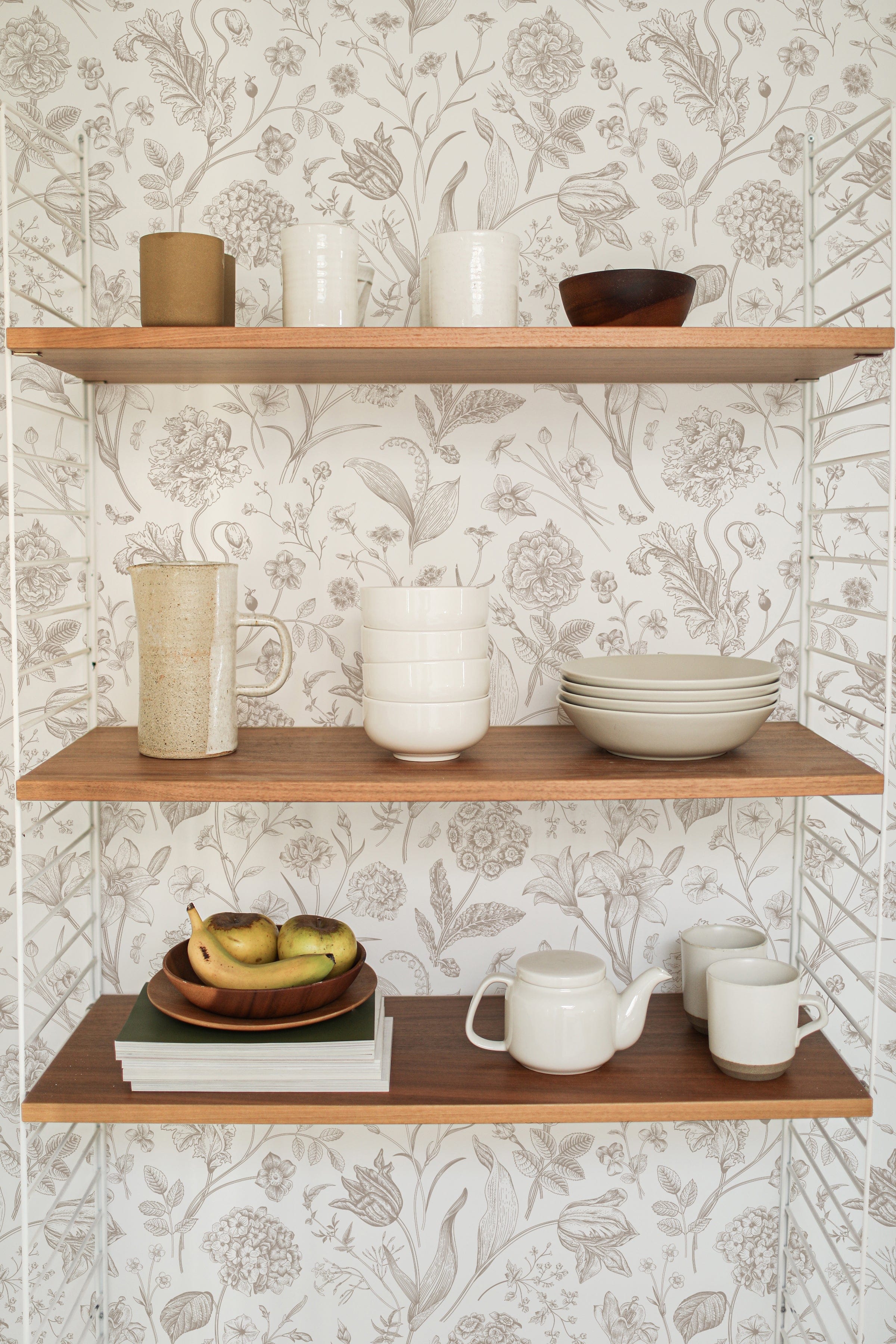 Open wooden shelves against a wall covered with Toile De Jouy Floral Wallpaper, which is adorned with delicate floral drawings. The shelves hold an array of kitchenware, adding a functional touch to the decorative space