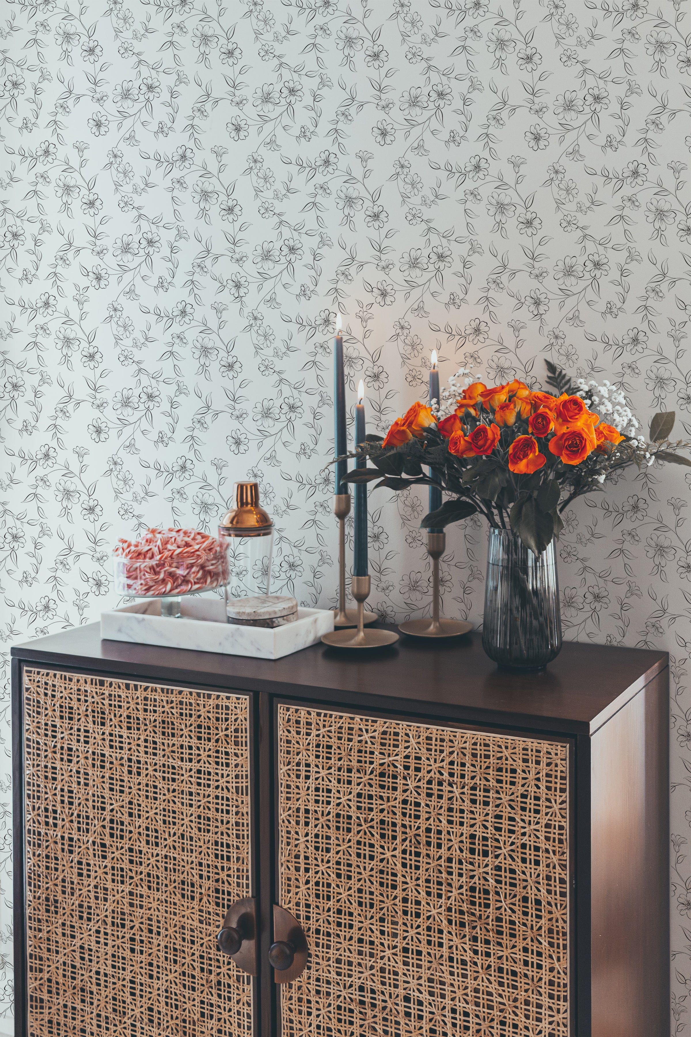 A stylish console table with decorative items against a wall covered in elegant monochrome wallpaper. The wallpaper features a delicate black and white floral pattern, adding a touch of sophistication to the space. The console table is adorned with a vase of orange roses, candles, and a tray, enhancing the elegant ambiance.