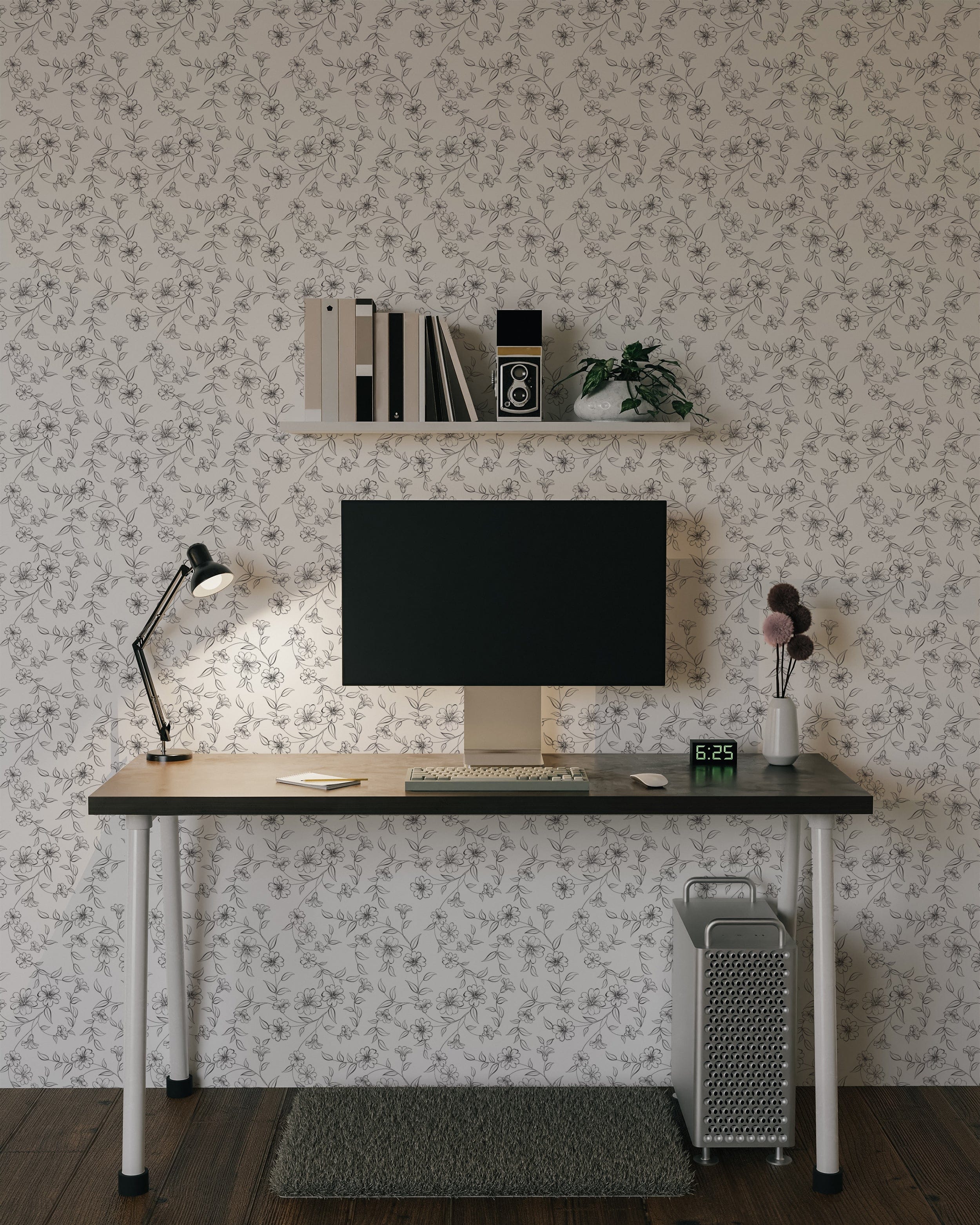 A modern workspace with a desk and computer set against a wall covered in elegant monochrome wallpaper. The wallpaper features a delicate black and white floral pattern, adding a touch of sophistication and style to the space. The desk is adorned with a lamp, a clock, and a vase, creating a clean and organized look