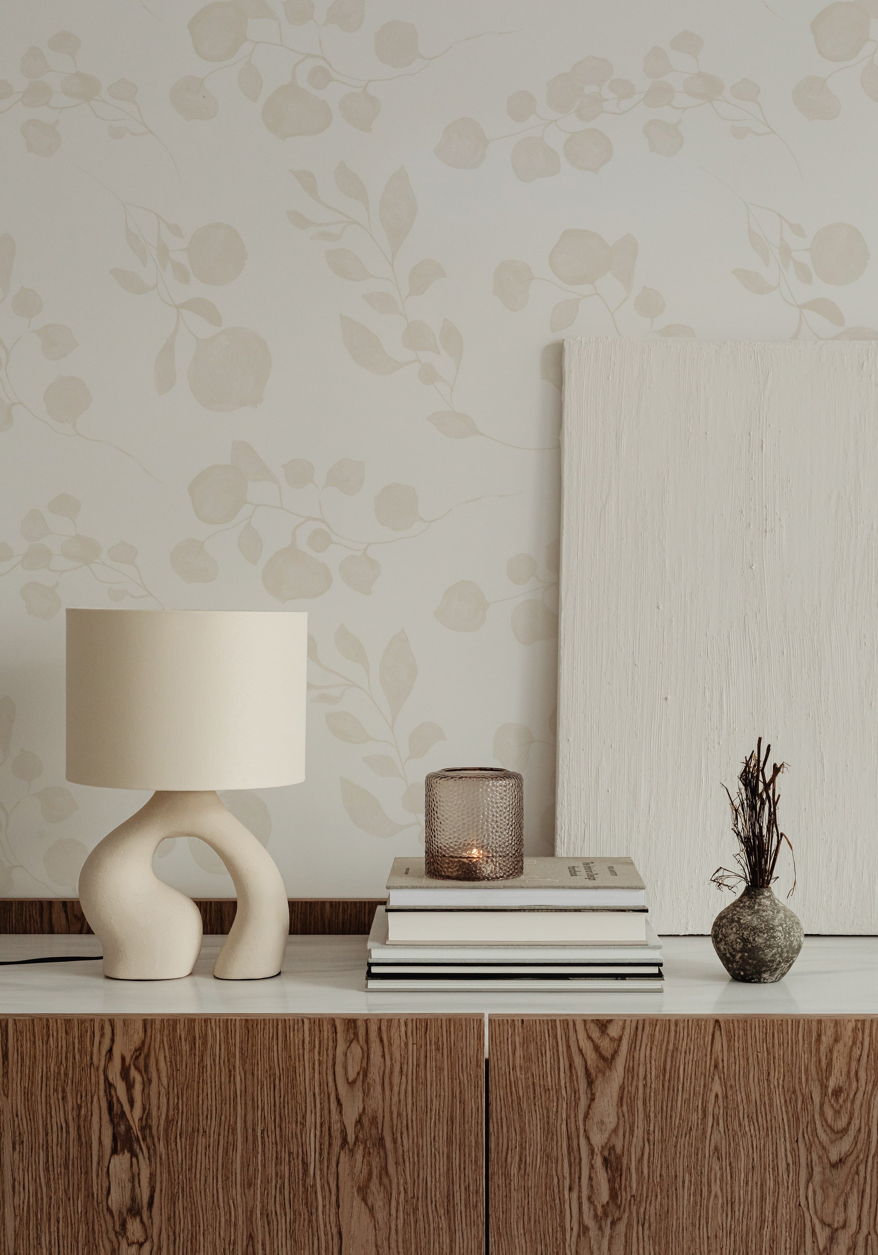 An elegantly styled room featuring the 'Subtle Botanique Wallpaper', with delicate beige botanical patterns over a light background, above a dark wood paneling. The decor includes a unique sculptural lamp on a wooden sideboard, adding to the room’s contemporary natural aesthetic.