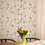 The Watercolour Daisy Wallpaper adds a delicate touch to a sunny dining area, complementing the natural wood furniture and a vase of fresh flowers on the table, which enhance the room's welcoming and cheerful ambiance.