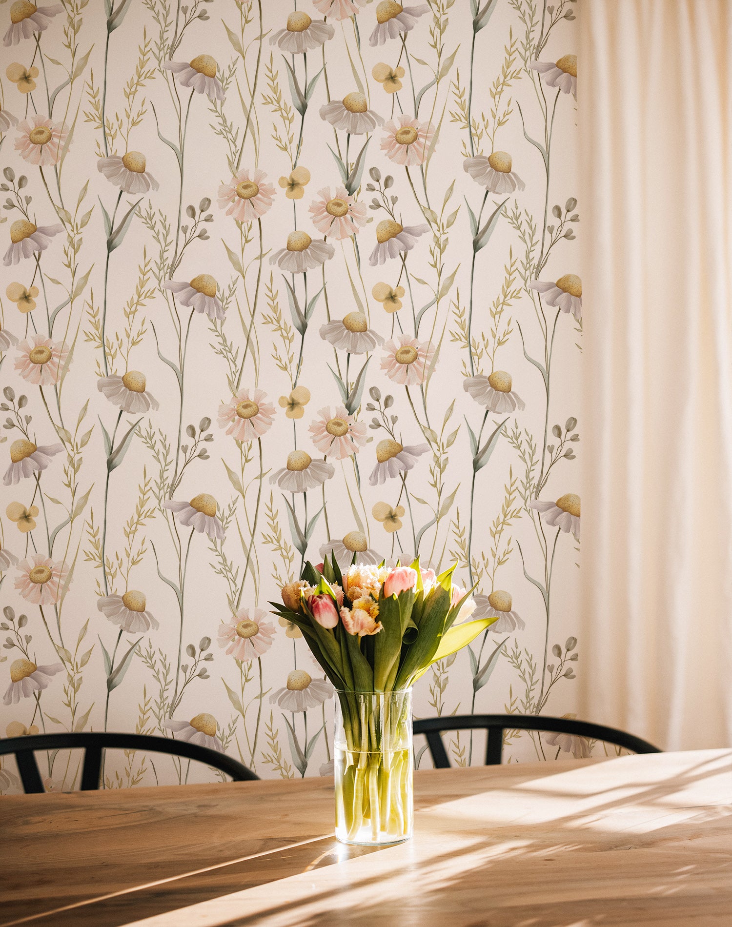The Watercolour Daisy Wallpaper adds a delicate touch to a sunny dining area, complementing the natural wood furniture and a vase of fresh flowers on the table, which enhance the room's welcoming and cheerful ambiance.