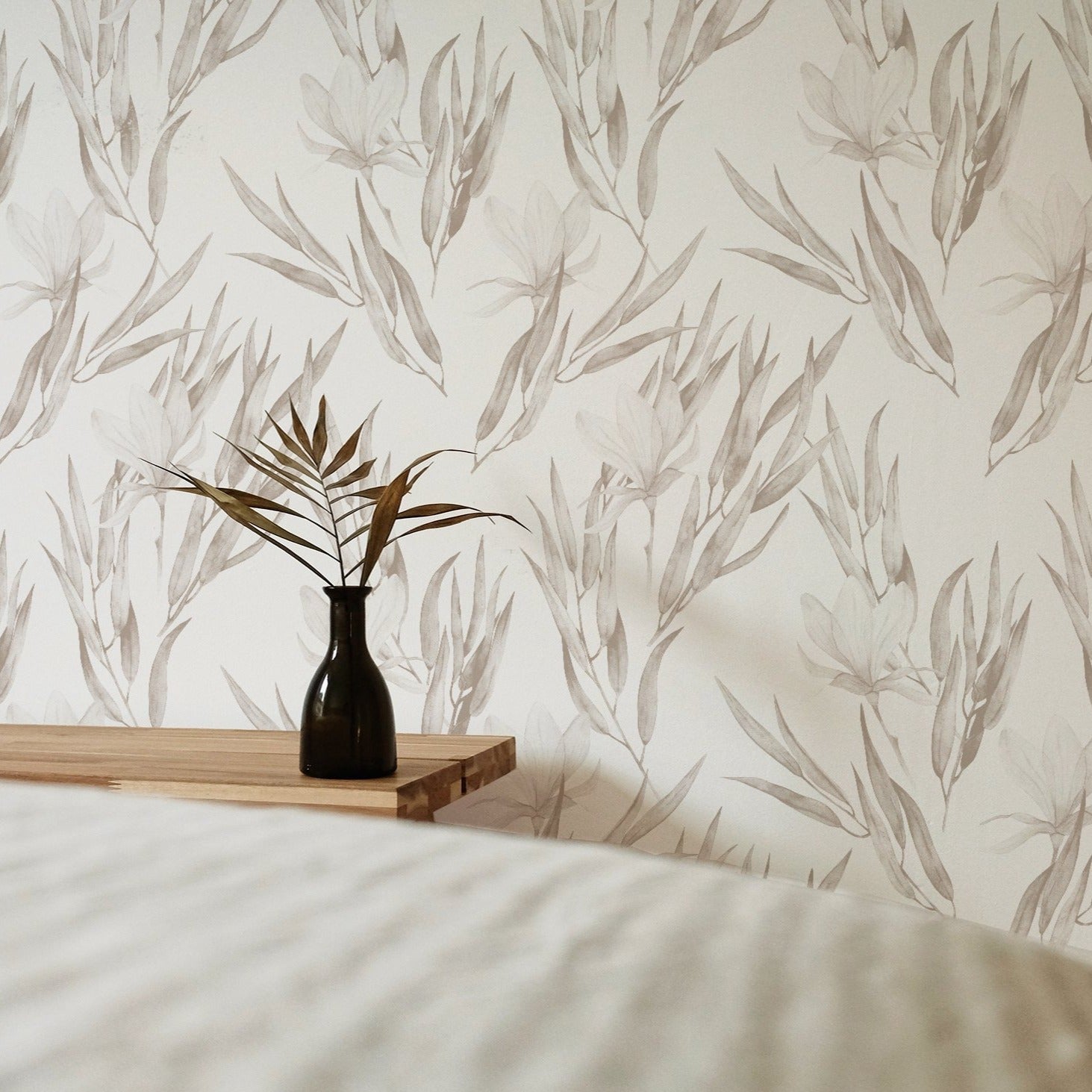 A serene bedroom decorated with wallpaper featuring soft watercolor illustrations of beige flowers and leaves. The space includes a wooden table with a dark vase holding dried foliage, enhancing the tranquil and elegant atmosphere.