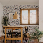 A cozy dining area with a wooden table and black chairs, featuring wallpaper with a muted terrazzo pattern. The space is adorned with a potted plant, wicker pendant light, and framed artwork, creating a warm and inviting atmosphere.