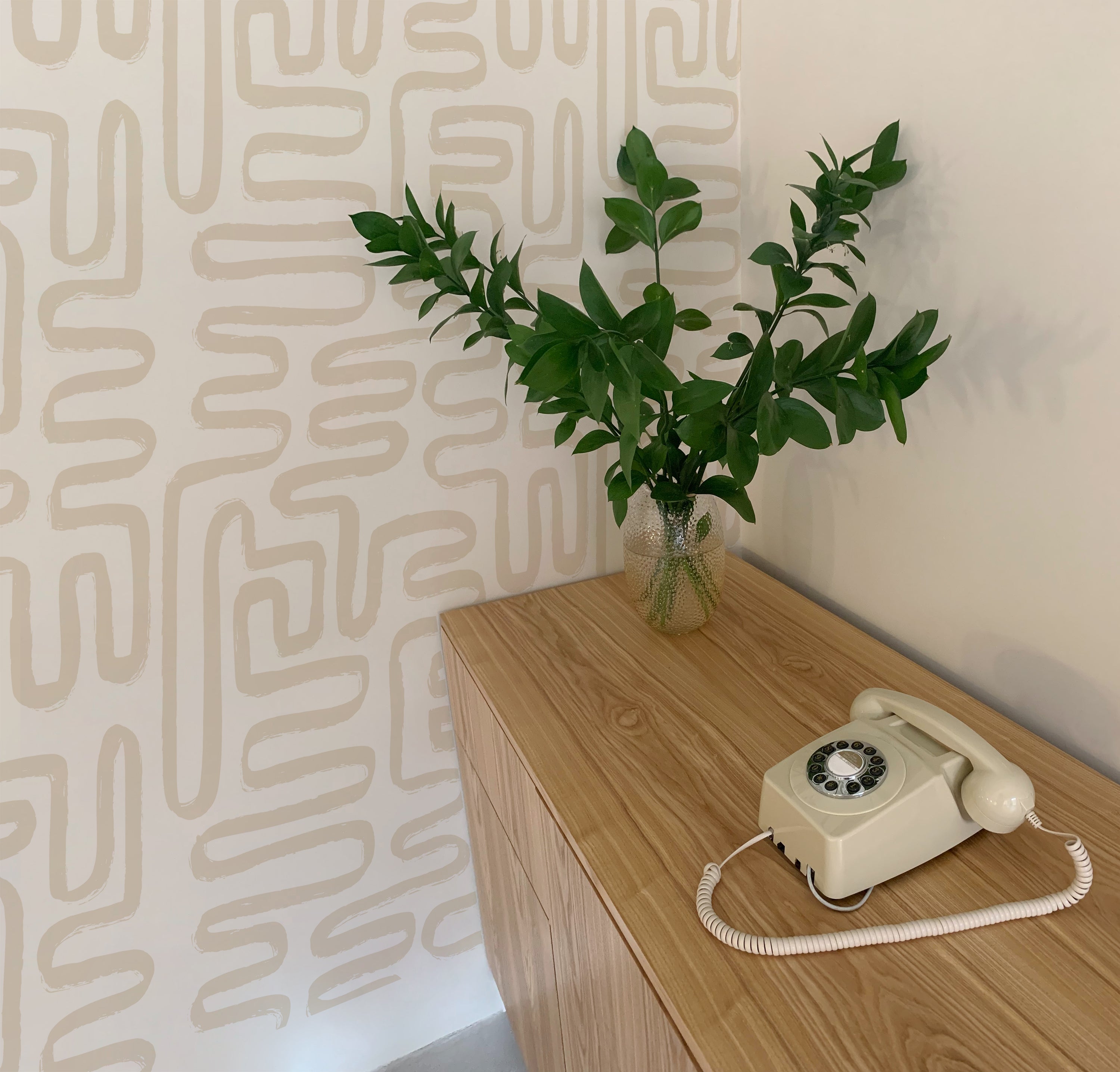 A snapshot of a modern, understated corner with clean lines, featuring the ecru geometric wallpaper. A vintage telephone on a light wood console table and a green leafy branch in a clear vase add a touch of retro charm.