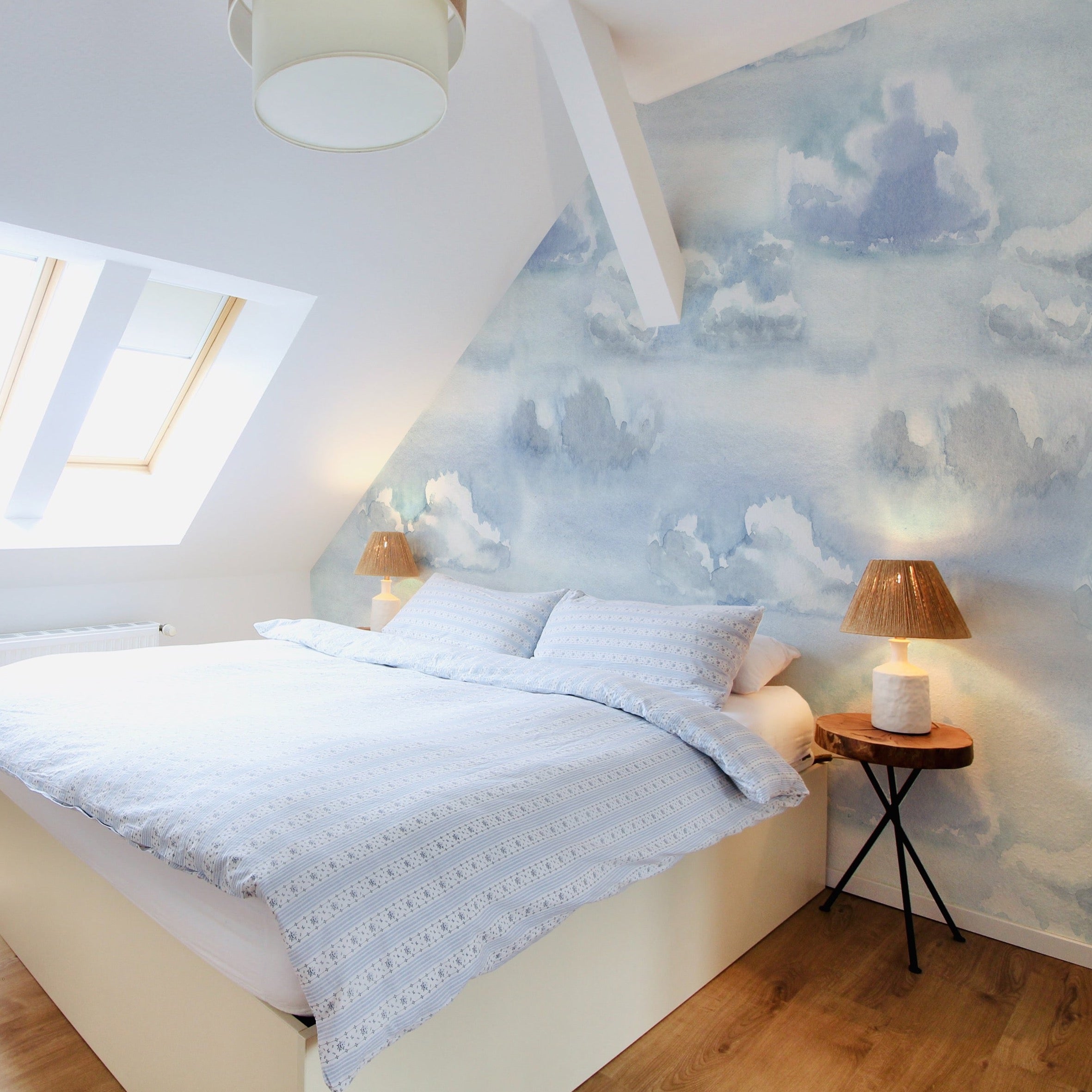 The Watercolour Cloud and Skies II wallpaper adorning a bedroom wall behind a neatly made bed, evoking a calm and dreamy atmosphere in the space.