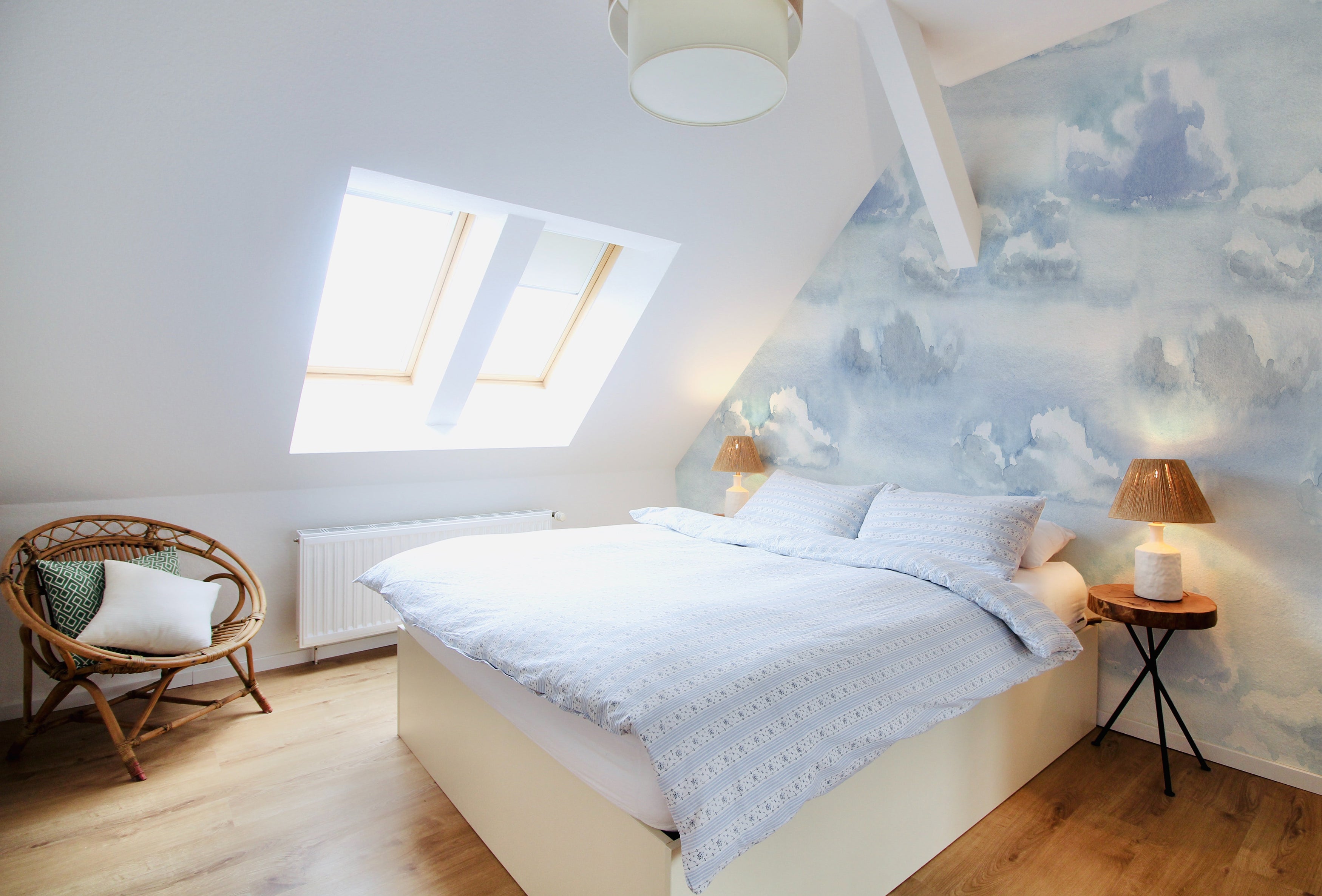 The Watercolour Cloud and Skies II wallpaper adorning a bedroom wall behind a neatly made bed, evoking a calm and dreamy atmosphere in the space.