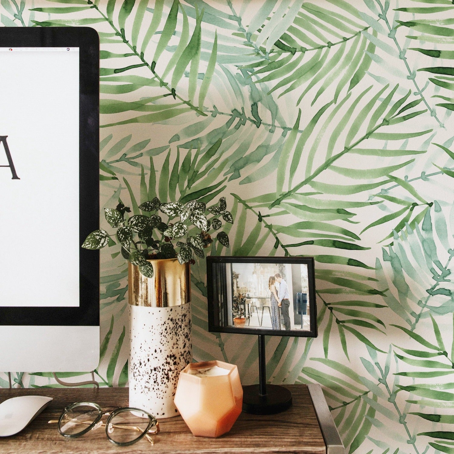 A cozy workspace is enlivened by the 'Hand Painted Tropical Wallpaper' featuring lush green watercolor palm leaves on a light background. The wallpaper brings a vibrant, organic feel to the area, complemented by a wooden shelf with a framed art print, books, and a modern computer setup with decorative vases and greenery.