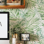 A cozy workspace is enlivened by the 'Hand Painted Tropical Wallpaper' featuring lush green watercolor palm leaves on a light background. The wallpaper brings a vibrant, organic feel to the area, complemented by a wooden shelf with a framed art print, books, and a modern computer setup with decorative vases and greenery.
