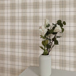 A subtle scene with a sleek white vase holding delicate flowers set against the Winter Plaid Wallpaper. The wallpaper's soft, neutral plaid pattern adds depth and a classic touch to the modern space, reflecting a blend of contemporary style and traditional comfort.